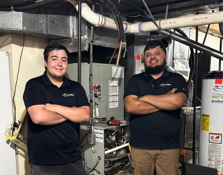 Two SwiftPro HVAC technicians standing arms crossed beside an open gas furnace and a water heater in a Northern Virginia basement